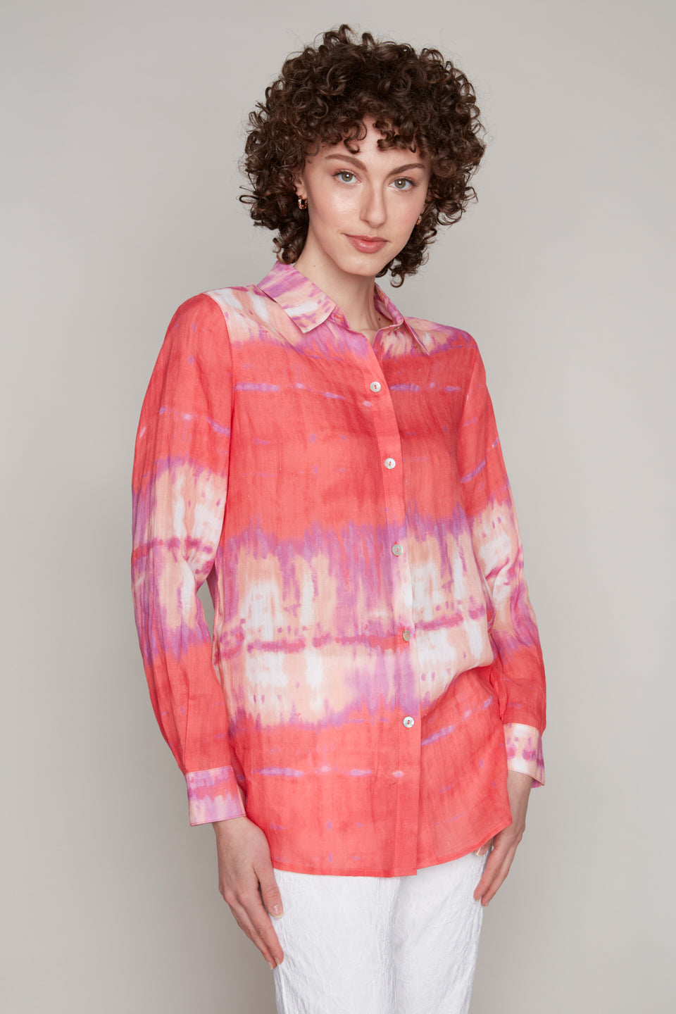 A fashion enthusiast stands gracefully against a plain background, wearing a Carre Noir Tie Dye Buttoned Blouse in bright pink, paired with crisp white pants.