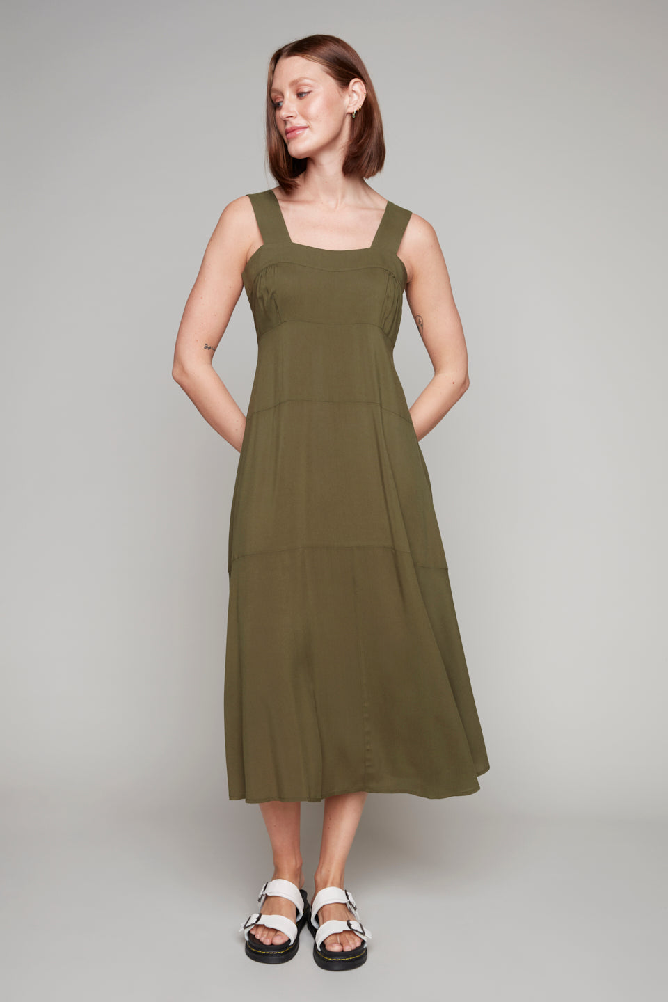 A person stands against a plain background wearing the Carre Noir Wide Strap Sleeveless Maxi in olive green, a true summer essential, paired with white sandals.