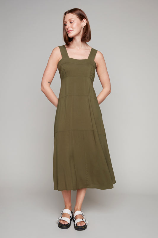 A person stands against a plain background wearing the Carre Noir Wide Strap Sleeveless Maxi in olive green, a true summer essential, paired with white sandals.