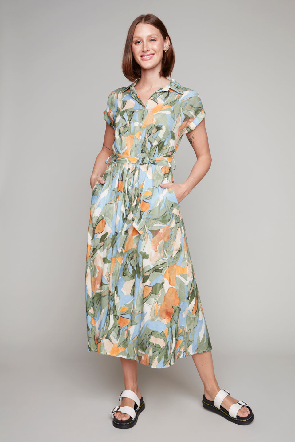 A smiling person stands against a plain backdrop, wearing the Carre Noir Short Sleeve Floral Dress with Pockets and a front waist tie, perfectly paired with white sandals.