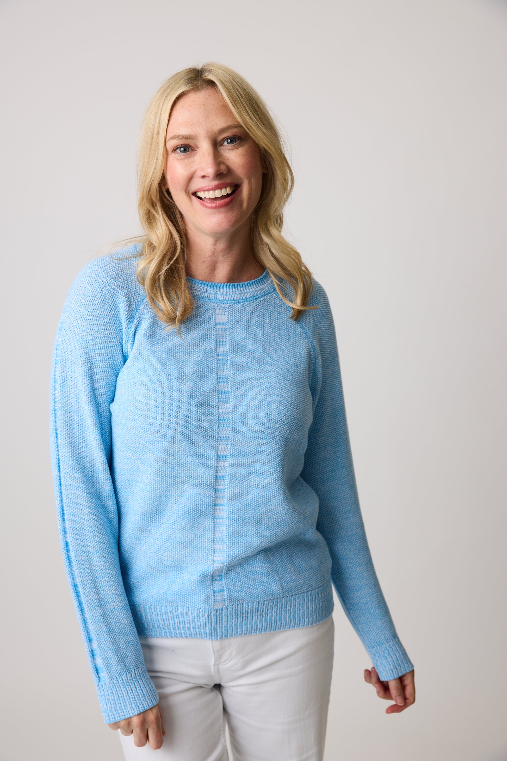 A long-haired blonde wearing the Cotton Country All Season Sweatshirt in light blue with white pants, smiles against a plain background. This versatile piece offers style and comfort for any occasion year-round.