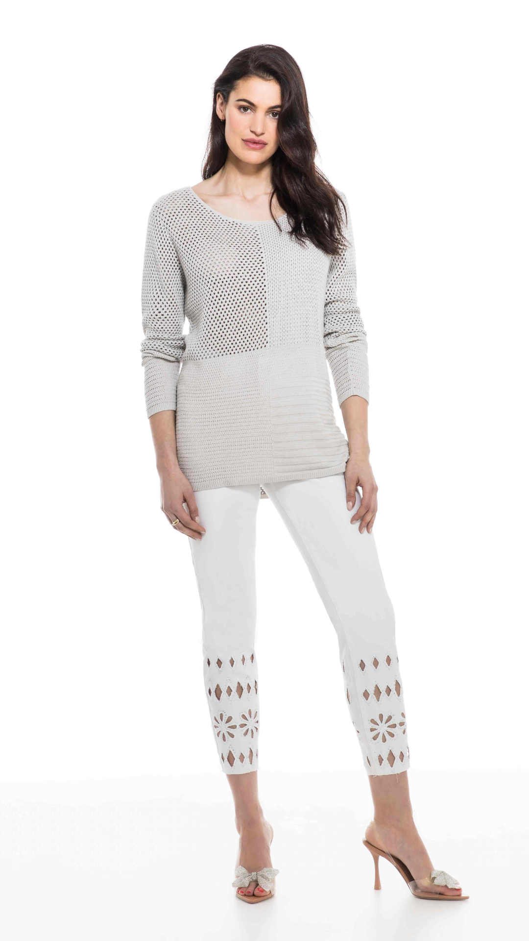 A woman stands confidently, wearing a casual, two-tone Orly Mesh Long Sleeve Sweater with white pants adorned with cut-out patterns, complemented by a pair of open-toe heels.