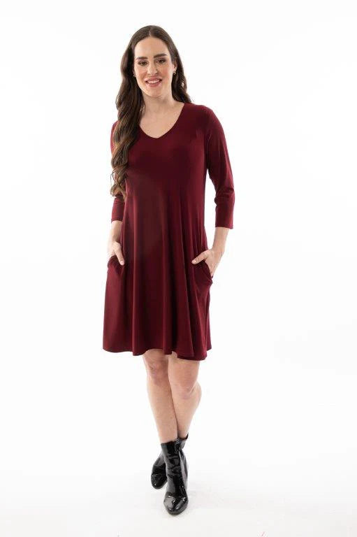 A woman in a Burgundy 3/4 Sleeve Dress with Pockets by Bali stands and smiles against a plain white background, her hands resting in the pockets of the knee-length dress.