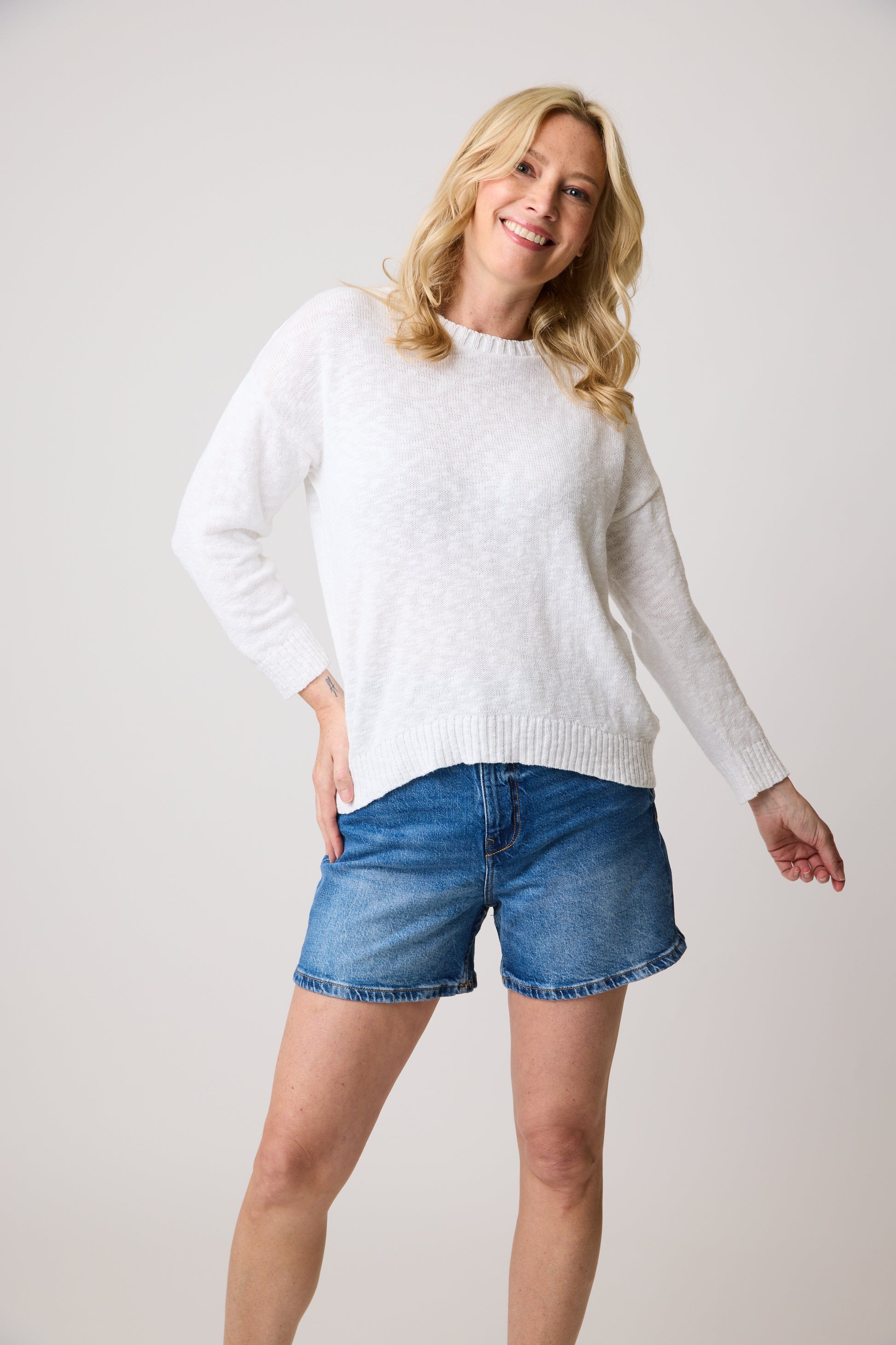 A blonde woman smiles in a Cotton Country Danbury Top—cozy and white—paired with denim shorts, against a plain white backdrop.