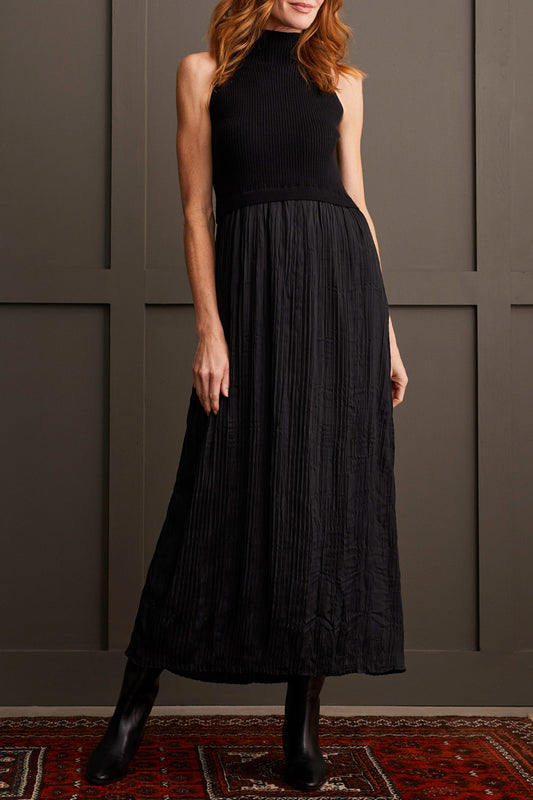 A person wearing Tribal's Sleeveless Twofer Maxi Dress, which features a high neck and a long, pleated skirt, stands against a gray paneled wall. They are also wearing black boots.