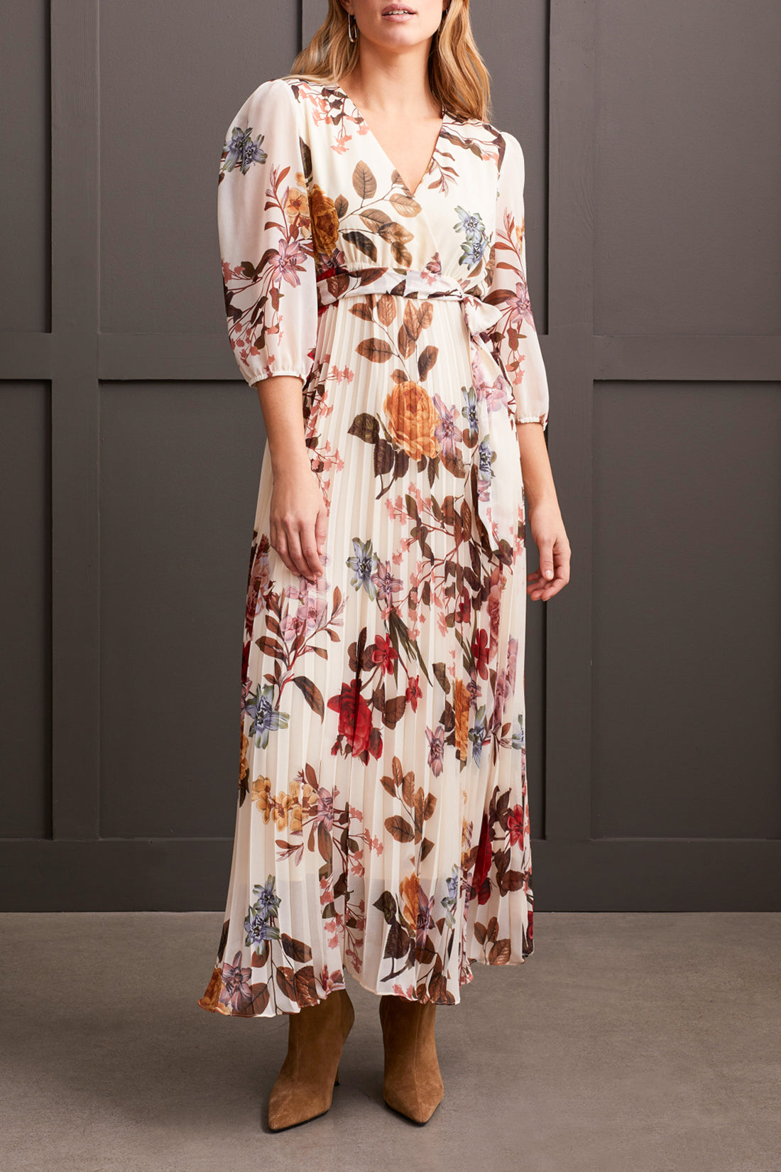 A person stands against a gray paneled background wearing Tribal's 3/4 Sleeve V Neck Dress with Side Tie, featuring floral detailing and a pleated design that accentuates the colorful flowers. The outfit is completed with tan boots.