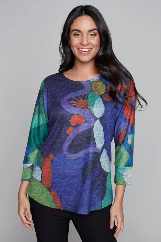 A person with long dark hair smiles while wearing the stylish and functional Round Neck Amazonia Top by Claire Desjardins, featuring colorful abstract shapes in blue, green, red, and purple tones. The long sleeves add a touch of sophistication to this trendy look.