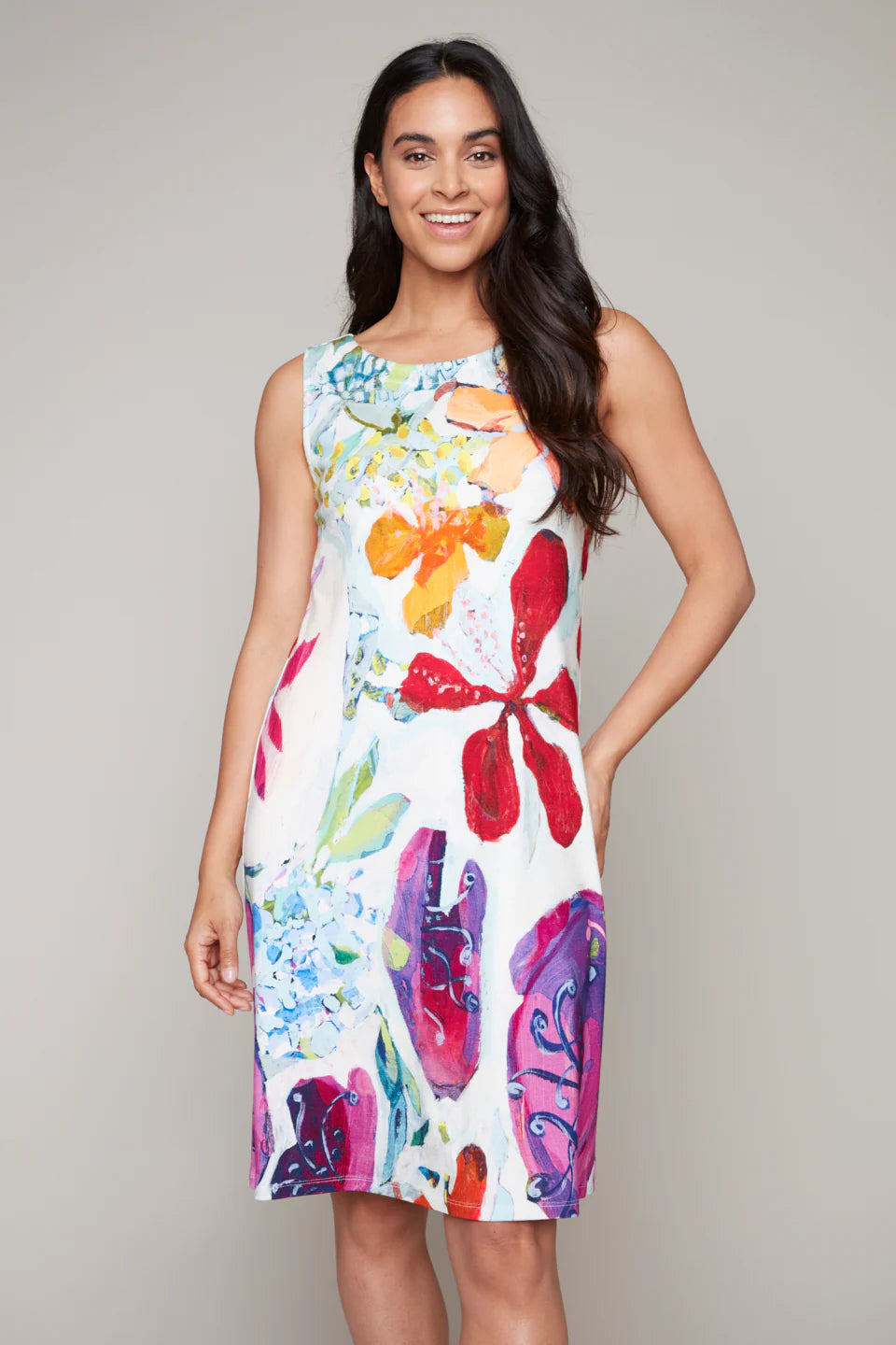 The woman is smiling in a Claire Desjardins Spring Sleeveless Dress with a floral pattern and flounce hemline, standing against a plain background.