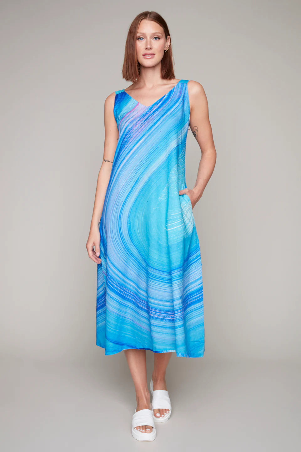 Against a plain backdrop, the person wears the Skate Park Sun Dress by Claire Desjardins, featuring a sleeveless, blue and turquoise swirl pattern ideal for outdoor activities, paired with white sandals.