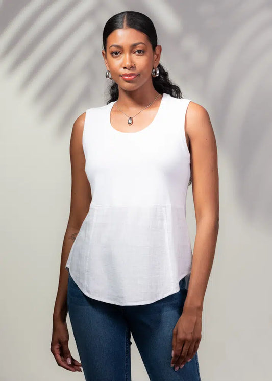 A woman in a sleeveless linen pullover top by Alison Sheri and blue jeans stands against a light background with leaf shadows, embodying the essence of summer days.