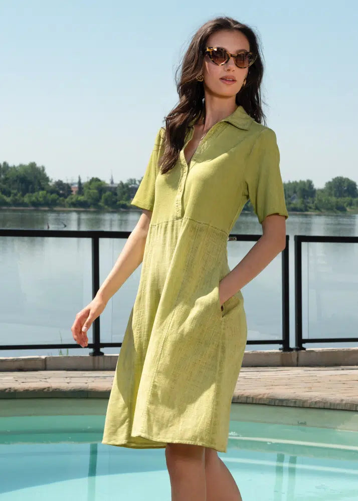 A woman in an Alison Sheri Button Front Linen Dress with Pockets and sunglasses stands by a pool, framed by glistening water and lush trees, ideal for any summer event.