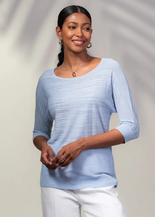 A woman beams joyfully in her light blue Alison Sheri Light Weight 3/4 Sleeve Sweater and white pants, epitomizing effortless fashion against a neutral backdrop.