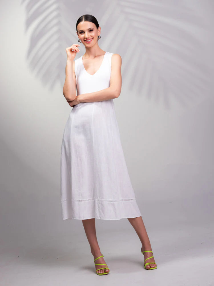 A woman in a white sleeveless Linen V-Neck Dress by Alison Sheri smiles while wearing green heels against a shadowed palm leaf backdrop—ideal for breezy summer days.