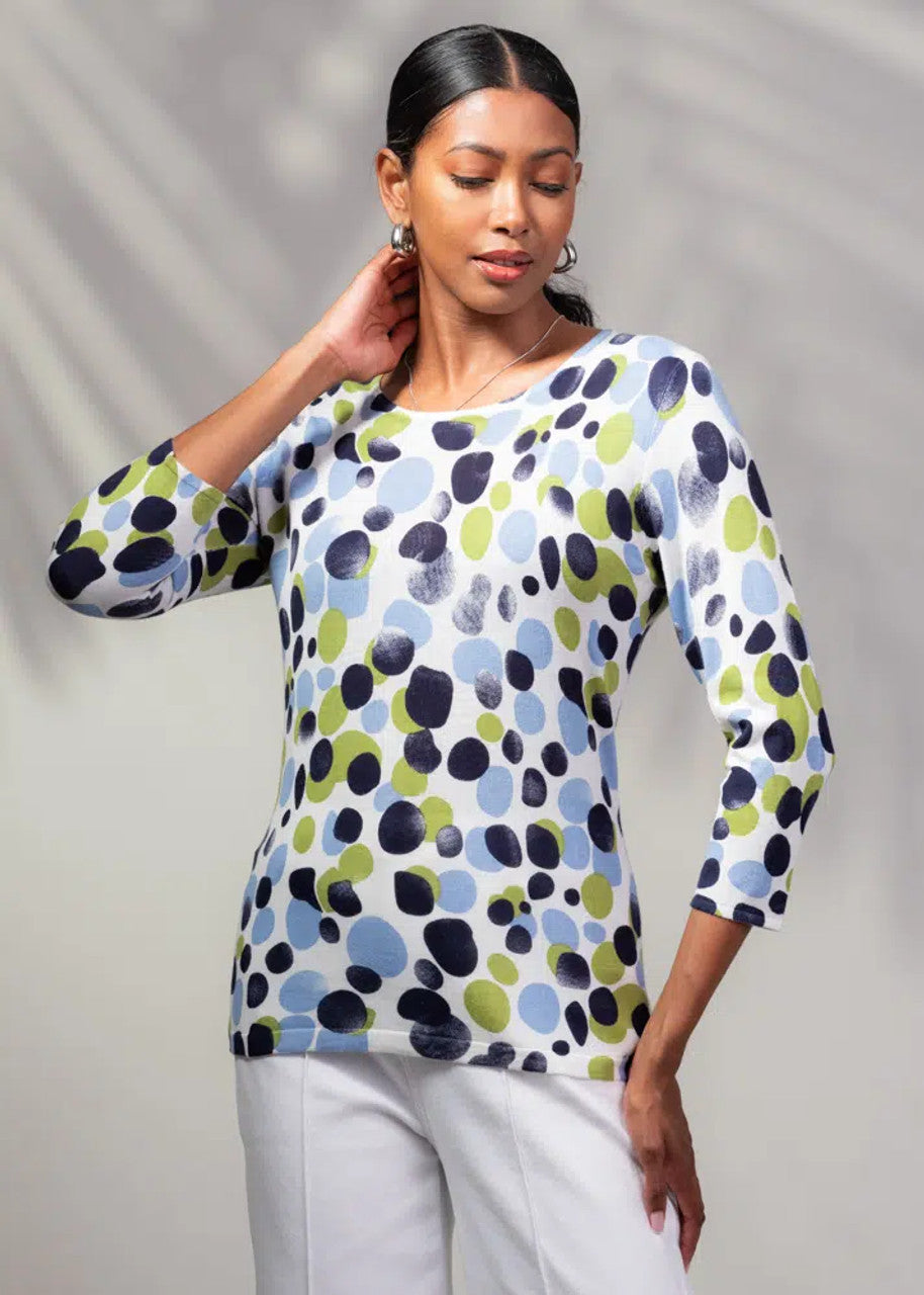 Wearing Alison Sheri's Dot Print Sweater featuring charming green, blue, and black spots, the person pairs it stylishly with white pants and stands gracefully with one hand on their neck against a softly blurred background.