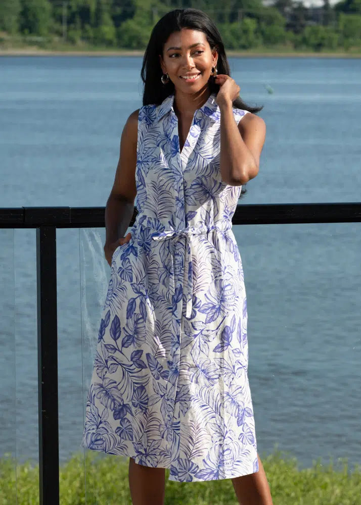 By the water, a woman wearing the Alison Sheri Button Up Floral Dress with Pockets in a blue and white pattern stands by a railing. She embraces summer style with its flattering fit, smiling warmly with one hand near her face.