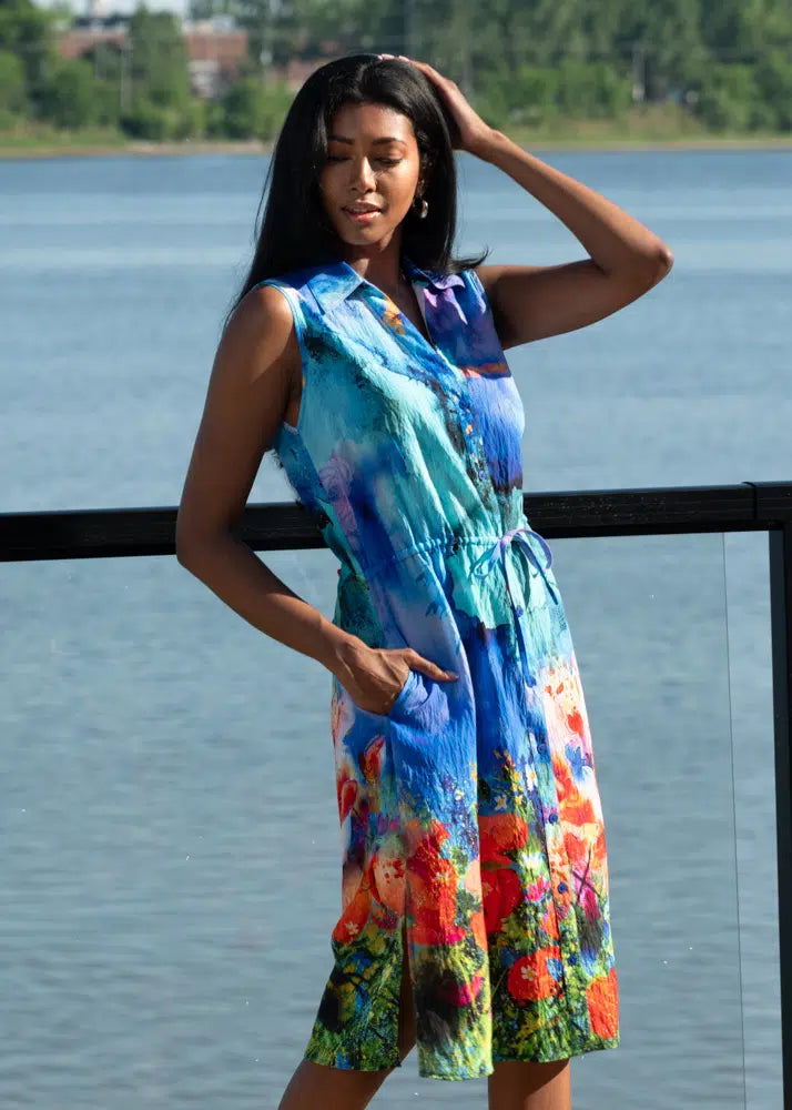 A woman in an Alison Sheri Floral Sleeveless Dress With Pockets stands by a railing with a river and trees behind her. She casually touches her hair with one hand while the other rests in her pocket, capturing the essence of a perfect summertime moment.