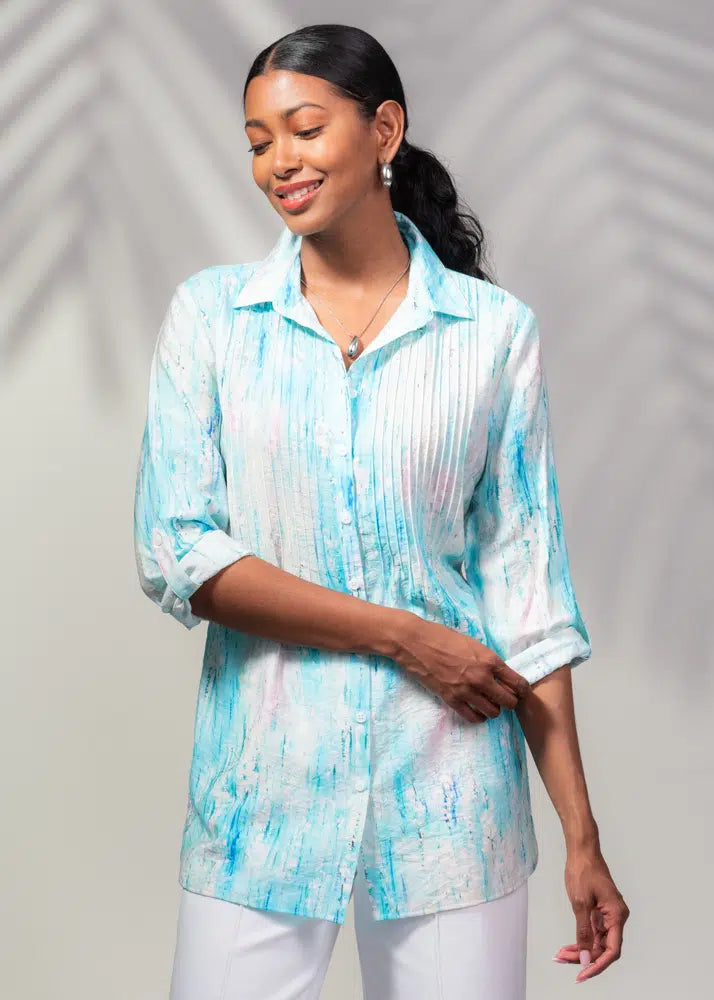 Smiling, a person models Alison Sheri's light blue 3/4 Sleeve Pleated Top with rolled-up sleeves and white pants against a shadowed background—an ideal summer wardrobe addition.