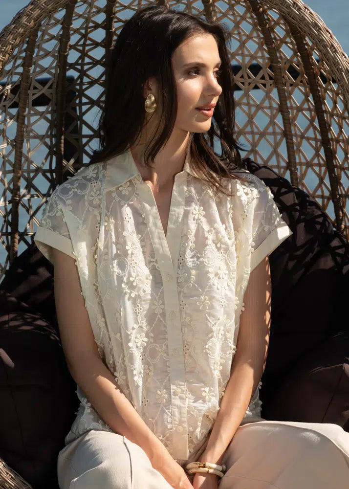 A woman sits on a wicker chair outdoors, wearing an Alison Sheri Embroidered Blouse with delicate floral details, gazing thoughtfully to her right.