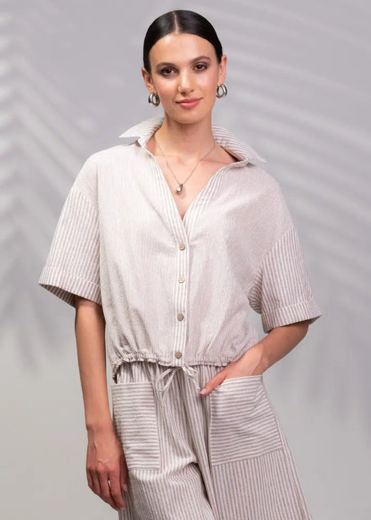 A woman wearing Alison Sheri's stylish striped short-sleeve button-up dress with a drawstring waist stands against a gray backdrop with leaf shadows—ideal for your summer wardrobe.