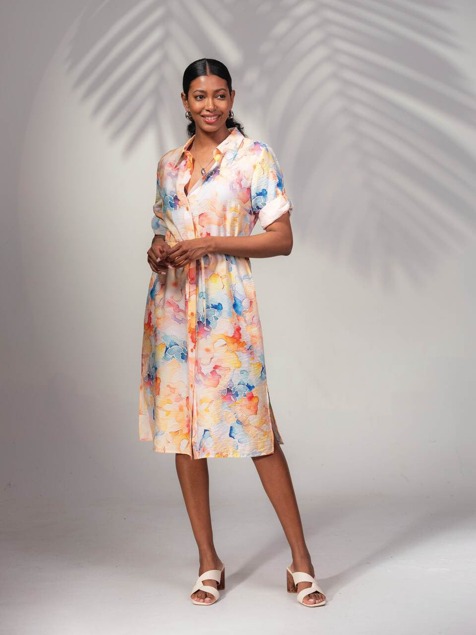 A woman in an Alison Sheri Floral Shirt Dress, bright and floral, smiles under palm-like fronds. Her white sandals enhance the summer's relaxed vibe as she poses effortlessly against a neutral backdrop.