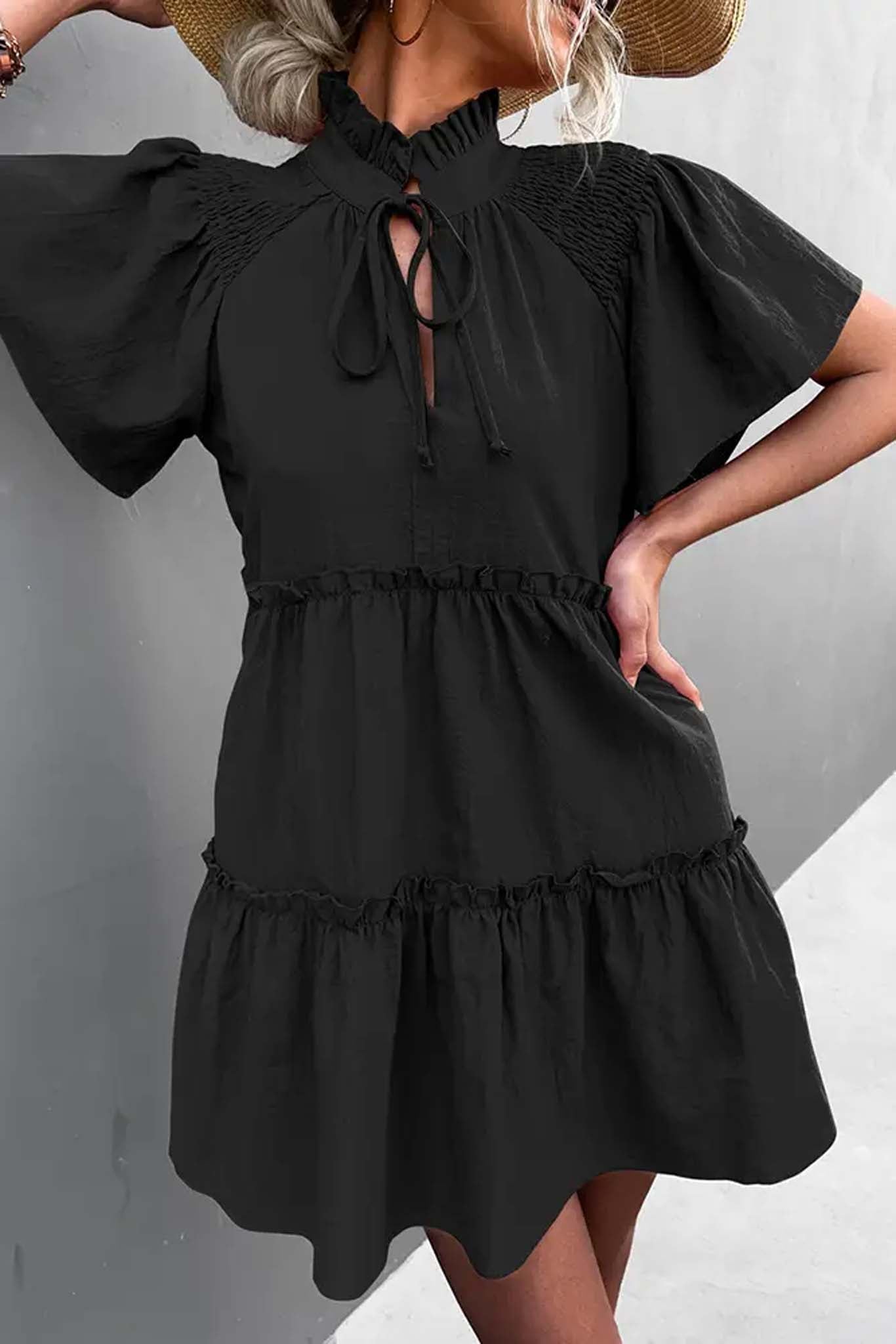 A woman in a black ruffled mini dress with puff sleeves and a tied neckline, holding the skirt, standing outdoors by Don't Be Chy Boutique.