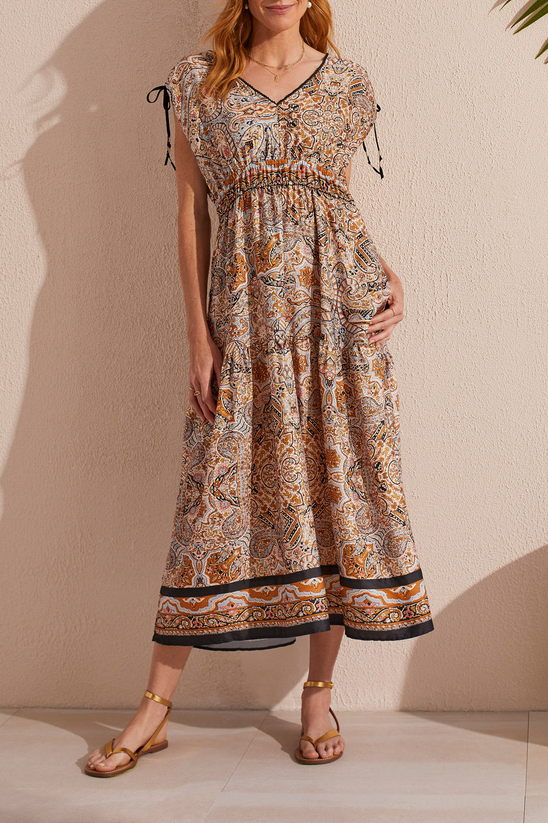 Woman posing in a Tribal Border Print Maxi Dress With Shoulder Ties and waist belt, accessorized with gold sandals.