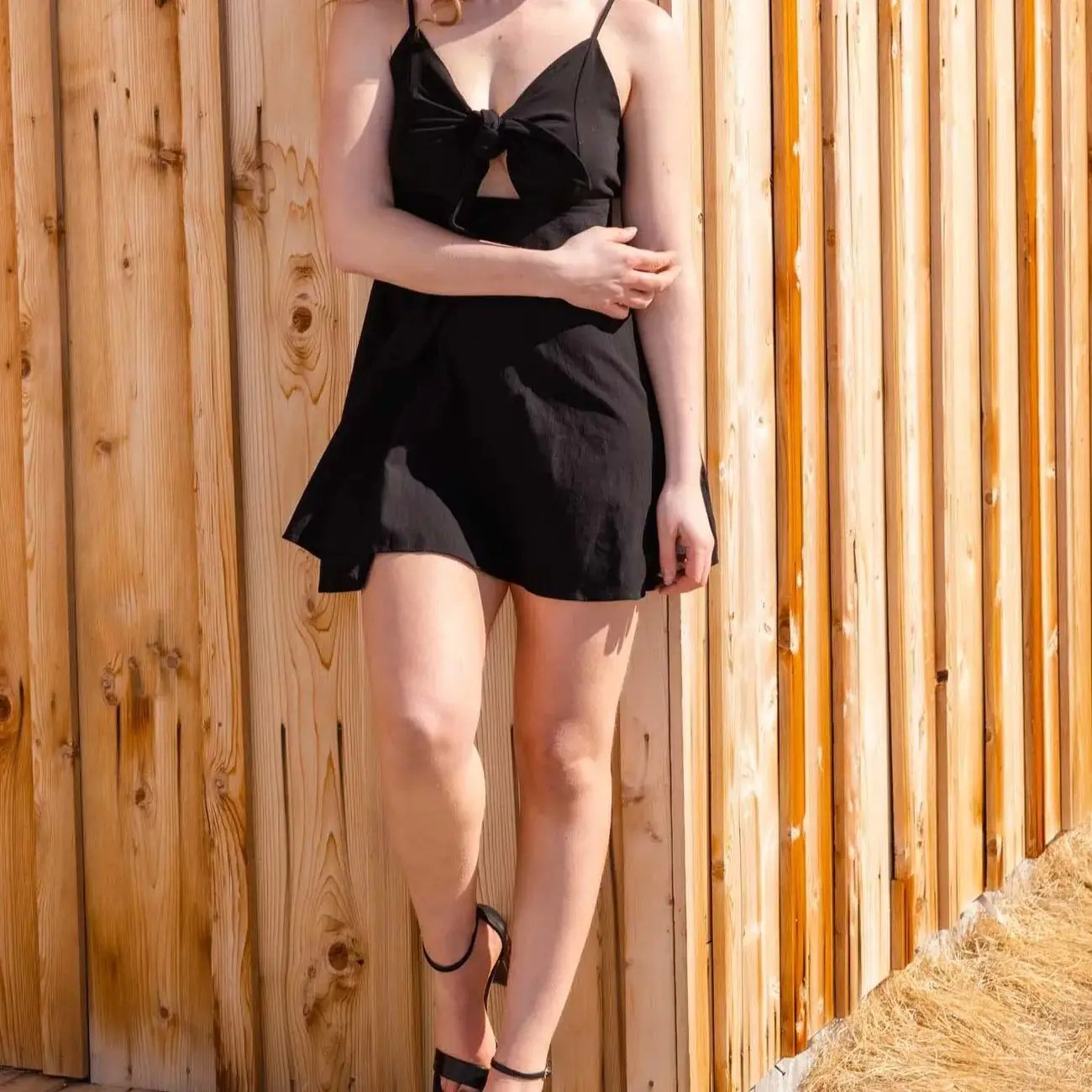 A woman wearing a Don't Be Chy Boutique Bow Knot Backless Sling Dress in black stands by a wooden fence, partially cropped to show from the neck down.