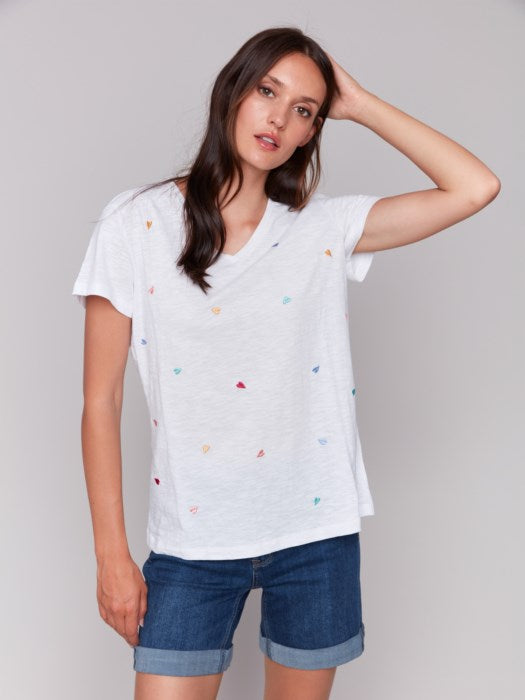 A person with long brown hair wears a Charlie B Short Sleeve V-Neck T-Shirt with Hearts Embroidery, looking down and smiling with arms gently crossed. The super soft fabric makes the shirt feel as cozy as it looks.