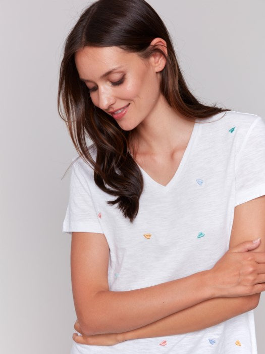 A person with long brown hair wears a Charlie B Short Sleeve V-Neck T-Shirt with Hearts Embroidery, looking down and smiling with arms gently crossed. The super soft fabric makes the shirt feel as cozy as it looks.