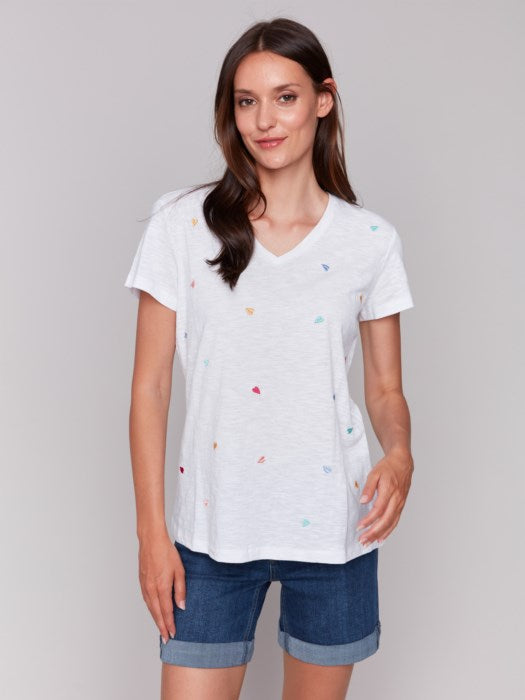 A person with long brown hair wears a Charlie B Short Sleeve V-Neck T-Shirt with Hearts Embroidery, looking down and smiling with arms gently crossed. The super soft fabric makes the shirt feel as cozy as it looks.