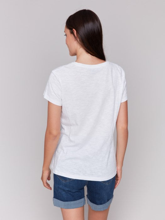 A person with long brown hair wears a Charlie B Short Sleeve V-Neck T-Shirt with Hearts Embroidery, looking down and smiling with arms gently crossed. The super soft fabric makes the shirt feel as cozy as it looks.