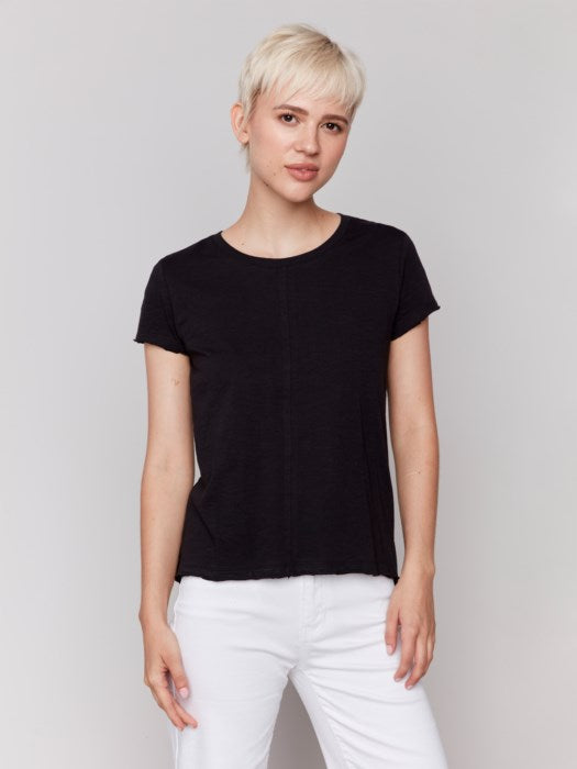 A person with short blond hair wears a black Charlie B Organic Cotton Slub Top and white organic cotton pants, standing against a plain background.