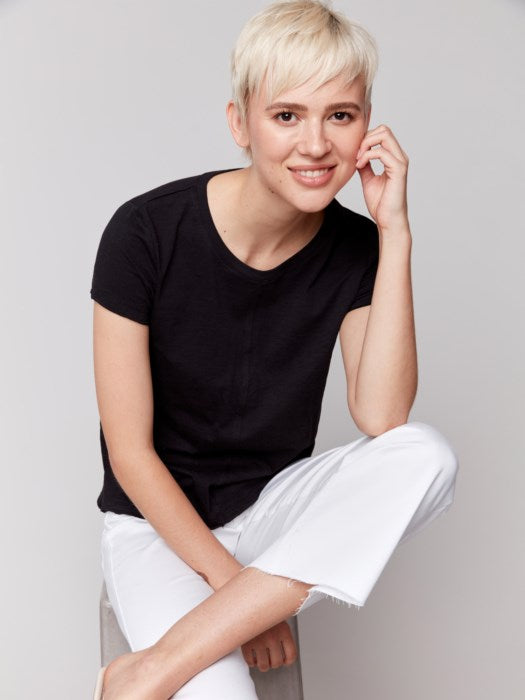 A person with short blond hair wears a black Charlie B Organic Cotton Slub Top and white organic cotton pants, standing against a plain background.