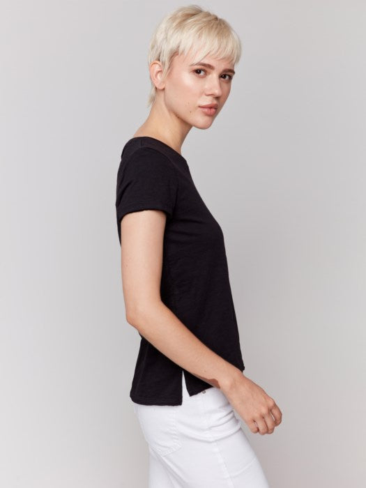 A person with short blond hair wears a black Charlie B Organic Cotton Slub Top and white organic cotton pants, standing against a plain background.