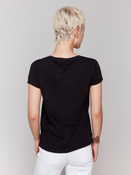 A person with short blond hair wears a black Charlie B Organic Cotton Slub Top and white organic cotton pants, standing against a plain background.