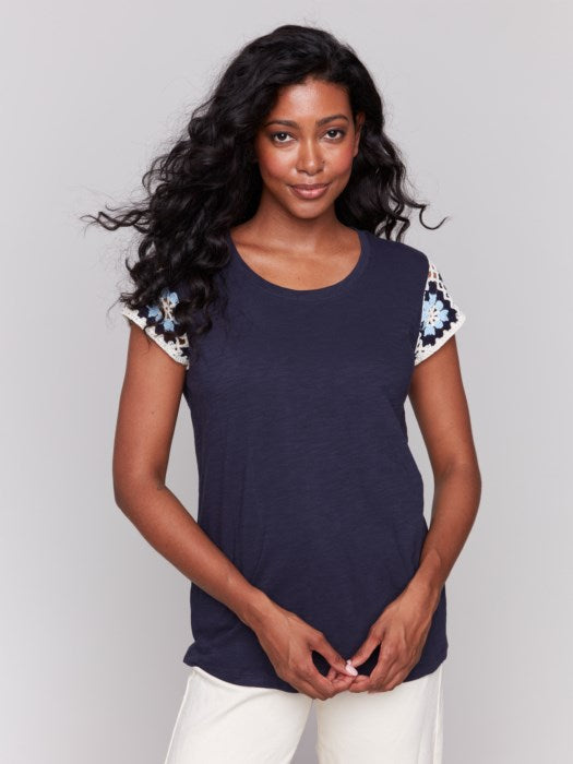 A person with long, wavy hair wears a navy Crochet Cap Sleeve T-Shirt by Charlie B, featuring floral-patterned short sleeves, paired with light-colored pants against a gray background.