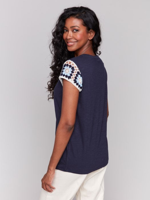 A person with long, wavy hair wears a navy Crochet Cap Sleeve T-Shirt by Charlie B, featuring floral-patterned short sleeves, paired with light-colored pants against a gray background.