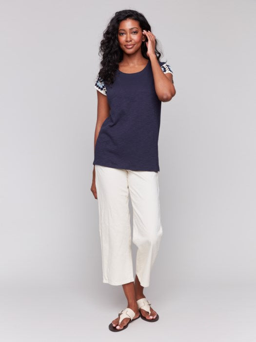 A person with long, wavy hair wears a navy Crochet Cap Sleeve T-Shirt by Charlie B, featuring floral-patterned short sleeves, paired with light-colored pants against a gray background.