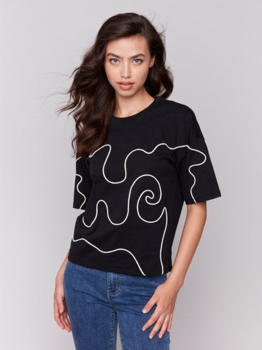 A woman with long hair wears a Charlie B Relaxed Fit Short Sleeve T-Shirt in black, showcasing white abstract line art, paired with blue jeans, ideal for a casual occasion, against a plain gray background.