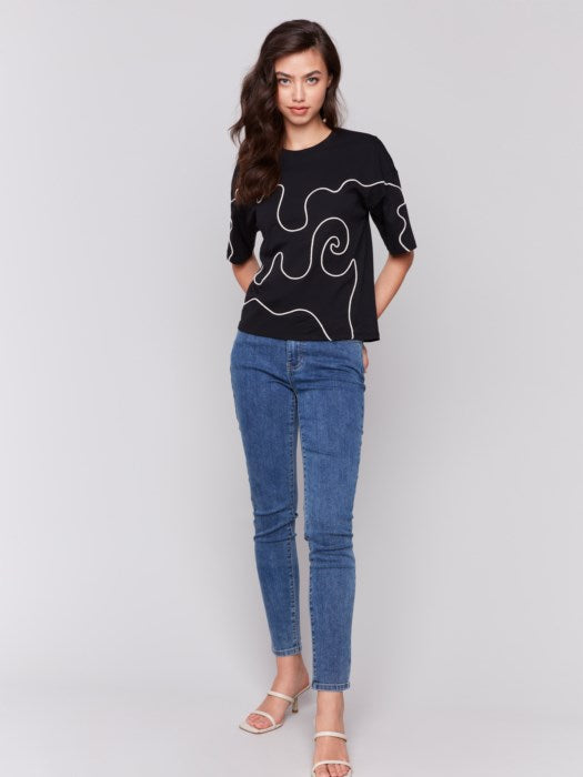 A woman with long hair wears a Charlie B Relaxed Fit Short Sleeve T-Shirt in black, showcasing white abstract line art, paired with blue jeans, ideal for a casual occasion, against a plain gray background.