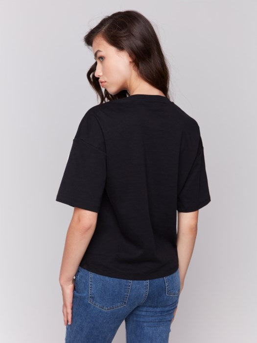 A woman with long hair wears a Charlie B Relaxed Fit Short Sleeve T-Shirt in black, showcasing white abstract line art, paired with blue jeans, ideal for a casual occasion, against a plain gray background.