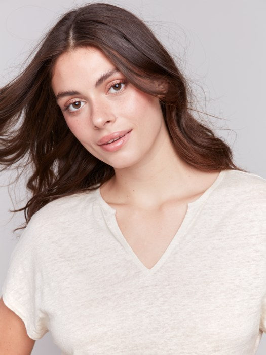 A person with long dark hair wears a stylish V-neck Foil Print Short Sleeve Dolman Top by Charlie B and white pants, posing against a plain background.