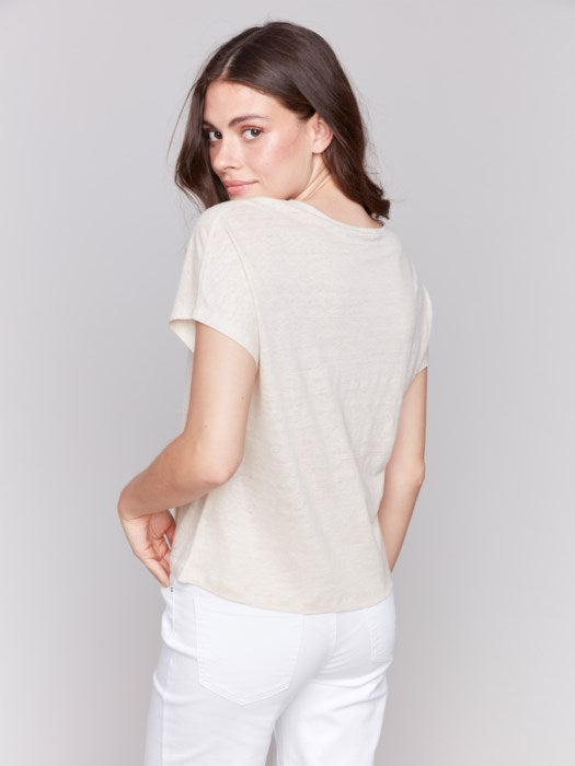 A person with long dark hair wears a stylish V-neck Foil Print Short Sleeve Dolman Top by Charlie B and white pants, posing against a plain background.