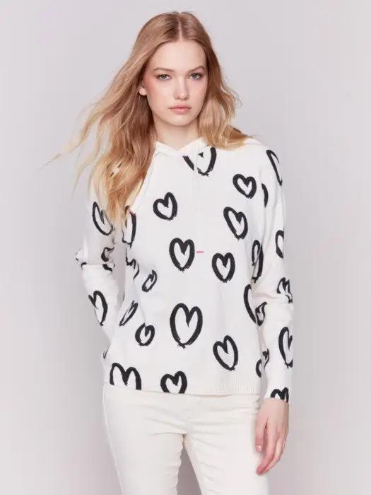 A person is standing against a plain background, facing sideways, wearing the Heart Printed Hoodie by Charlie B, featuring a cute design, along with white pants.