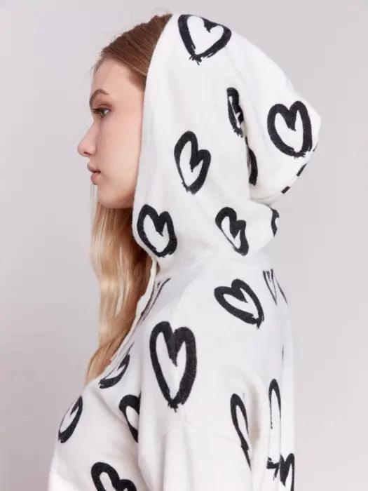 A person is standing against a plain background, facing sideways, wearing the Heart Printed Hoodie by Charlie B, featuring a cute design, along with white pants.