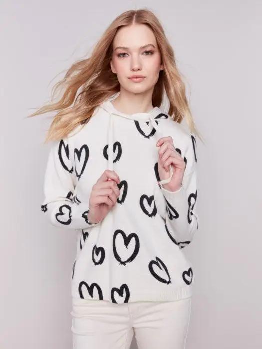 A person is standing against a plain background, facing sideways, wearing the Heart Printed Hoodie by Charlie B, featuring a cute design, along with white pants.