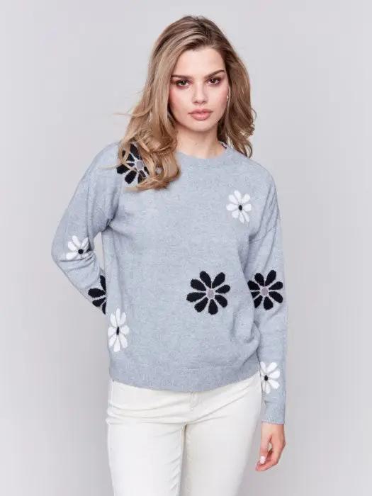 A fashionable woman with wavy blonde hair wears a cozy, light blue Printed Crew Neck Pullover Sweater by Charlie B, featuring black and white floral decorations, as she stands against a plain background.