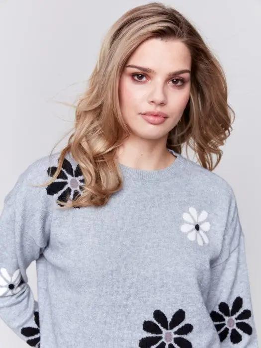 A fashionable woman with wavy blonde hair wears a cozy, light blue Printed Crew Neck Pullover Sweater by Charlie B, featuring black and white floral decorations, as she stands against a plain background.