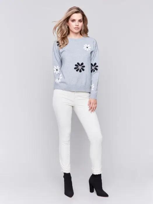 A fashionable woman with wavy blonde hair wears a cozy, light blue Printed Crew Neck Pullover Sweater by Charlie B, featuring black and white floral decorations, as she stands against a plain background.