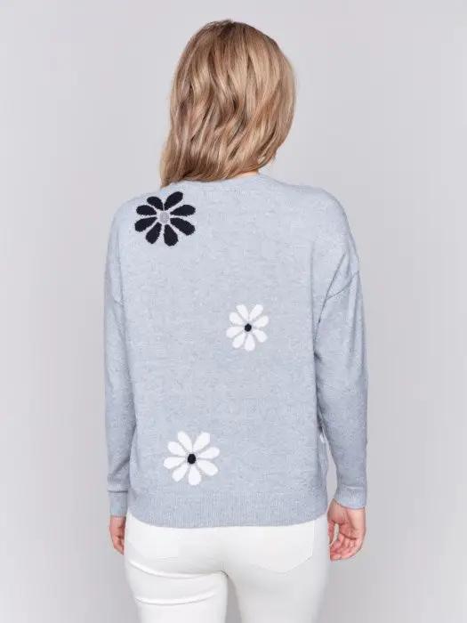 A fashionable woman with wavy blonde hair wears a cozy, light blue Printed Crew Neck Pullover Sweater by Charlie B, featuring black and white floral decorations, as she stands against a plain background.